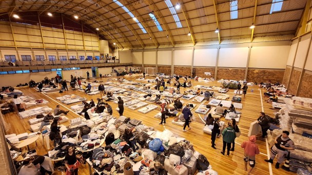Cete recebendo desalojados de Porto Alegre devido a enchentes. Imagem aberta e do alto do interior do ginásio, no qual há diversas pessoas, além de objetos e colchões espalhados pelo chão.